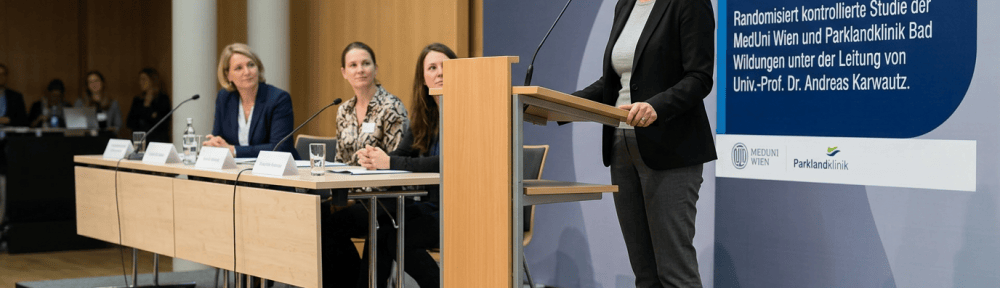 Speaker at podium beside poster reading: Begleitetes Selbstlernprogramm für Frauen und Mädchen mit Bulimia Nervosa. Randomisiert kontrollierte Studie der MedUni Wien und Parklandklinik Bad Wildungen unter der Leitung von Univ.-Prof. Dr. Andreas Karwautz. MedUni Wien. Parklandklinik.